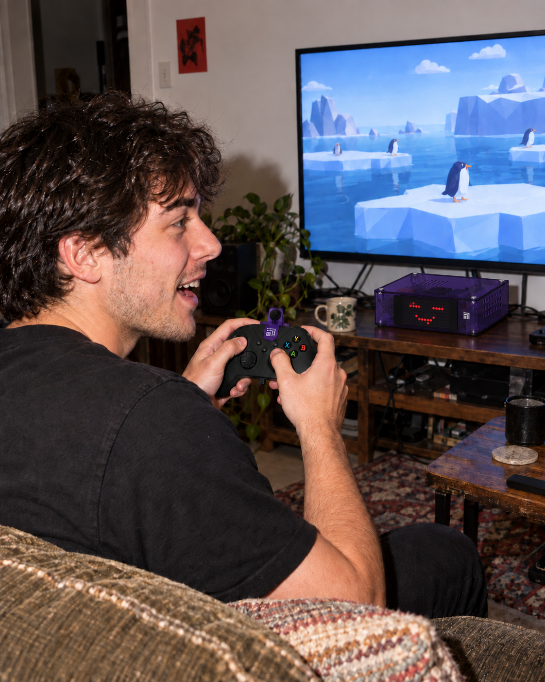 A person on a couch holds a controller with a purple microphone near their face, looking toward a penguin game on TV and the Threebox beneath it.